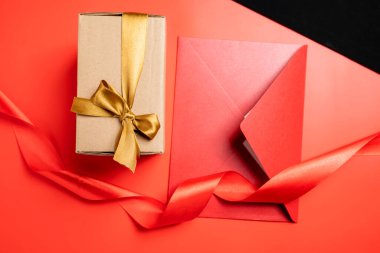 Altın kurdeleli kahverengi bir hediye kutusunun üst görüntüsü ve renkli bir arka planda kırmızı bir zarf. Model ya da kopyalama alanı. Yıldönümleri, kutlamalar, hediyeler veya sürprizler için dekorasyon kavramı