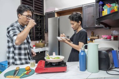 İki Endonezyalı güneydoğu Asyalı adam mutfakta birlikte yemek pişiriyor. İçkiyi tatmak ve gaz ocağında tavada yemek pişirmek. Yemek pişirme etkinliği