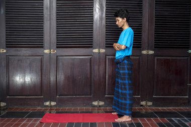 Mavi gömlekli Müslüman adam ahşap bir kapının önünde dua ediyor. Mavi bir etek giyiyor ve birini bekliyor.