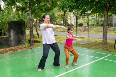 Bir kadın ve bir çocuk yeşil kortta yoga yapıyorlar. Kadın çocuğa nasıl poz verileceğini öğretiyor.