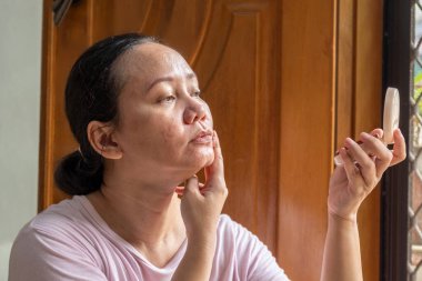 Kadın aynada kendine bakıyor. Makyaj yapıyor. Sahne ciddi ve odaklı.