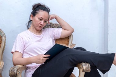 Kadın bir sandalyede oturmuş kitap okuyor. O, pembe bir gömlek ve siyah pantolon giyiyor. Sahne sakin ve huzurlu. Kadın kitabına odaklanmış.