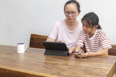 Endonezyalı kadın ve kızı evde birlikte bir proje üzerinde çalışıyorlar. Kadın çocuğun tableti kullanmasına yardım ediyor.