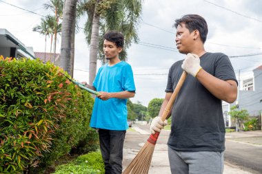 İki Endonezyalı Güneydoğu Asyalı bahçıvan çiti makaslarla buduyor, diğeri ise arka bahçede süpürgeyle süpürürken. Bahçe işleri.