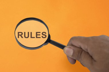 Bir el tarafından tutulan büyüteç, turuncu arka planda gösterilen kurallara odaklanıyor. Kurallar, düzenlemeler ve iş hayatına uyum kavramı