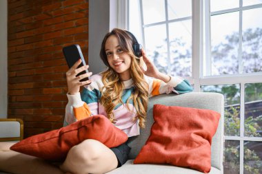 Endonezyalı güneydoğu Asyalı kadın evinde bir kanepede oturuyor, kulaklıkla müzik dinliyor ve elinde cep telefonu tutuyor, gülümsüyor ve rahatlıyor.