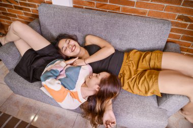 Endonezyalı güneydoğu Asyalı kadınlar modern bir iç mekanda rahat gri bir kanepede dinleniyorlar. Gülüyorlar ve gülümsüyorlar, birlikte hoş ve parlak bir oturma odasında vakit geçiriyorlar.