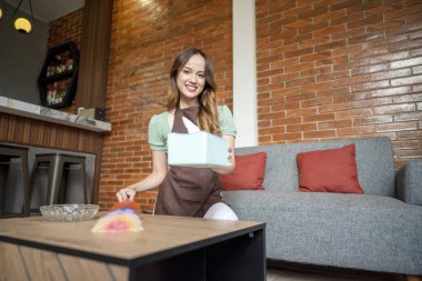 Endonezyalı güneydoğu Asyalı kadın ahşap bir masayı temizlemek için tüy tozu kullanıyor. Yanında da mendil kutusu ve cam kül tablası var. Temizlik ve temizlik.