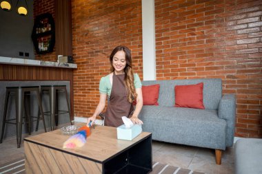 Endonezyalı güneydoğu Asyalı kadın ahşap bir masayı temizlemek için tüy tozu kullanıyor. Yanında da mendil kutusu ve cam kül tablası var. Temizlik ve temizlik.