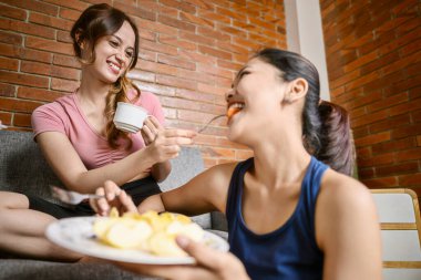 Endonezyalı Güneydoğu Asyalı kadınlar idmandan sonra içeride meyve salatası yiyorlar. Birlikte oturma odasında oturuyorlar, sağlıklı bir yemek yiyorlar ve gülümsüyorlar.