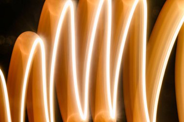 Akan ışık izlerini içeren soyut bir görüntü, sıcak ve parlak bir tonla kıvrımlı çizgiler oluşturuyor. Işık, organik ve dinamik bir dokuyu karanlık bir arkaplana boyar.