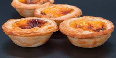 Koyu gri arka planda dört küçük kremalı tart. Öndeki iki tarta özenli odaklanır ve görünüm yoluyla yumuşar.