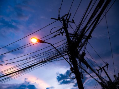 Silhouette of Electric Power Pole with Complex Cable Network Against Dramatic Blue Twilight Sky with Street Light
