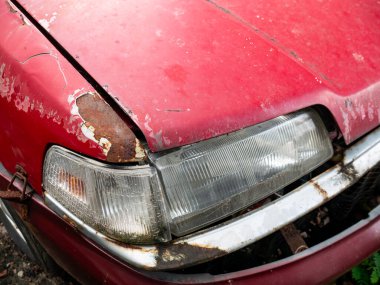 Kırmızı arabanın kaputunda paslanma ve soyulmuş boya hasarı var. Ön farın üzerinde metal bozulması var.