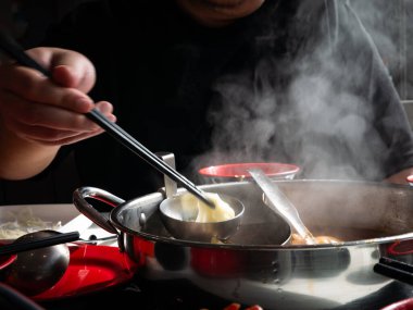 Yemek çubuklarıyla shabu shabu yiyen ve restoran masasındaki metal tencerede et suyu tüttüren biri.