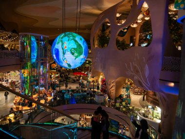 Bangkok 'taki Siyam Paragon' un avlusunu müşteriler ve lokantalar doldururken, büyük, aydınlatılmış bir dijital küre görüntüsü yer alıyor. Sahne, şehrin üst düzey perakende satış ve modern yaşam tarzını yansıtıyor..