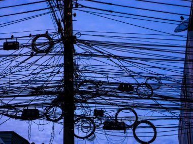Yoğun elektrik kabloları ve kablolar ahşap bir direğin etrafında yoğun bir siluet oluşturuyor. Derin mavi bir gökyüzüne karşı, kentsel elektrik altyapısı karmaşıklığı..