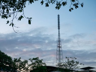 Uzun, metalik kafes iletişim kulesi, çevreleyen ağaç dalları ve yaprakları, teknoloji ve doğa çatışmaları ile dramatik bir akşam gökyüzüne ev sahipliği yapıyor..