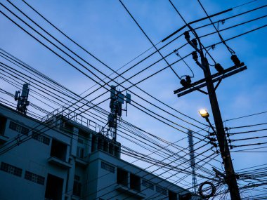 Arka planda haberleşme kuleleri olan bulutlu bir gökyüzüne karşı siluetlenmiş küçük bir ampulle ahşap elektrik direğinin üzerindeki karmaşık elektrik kabloları ve kablolar..