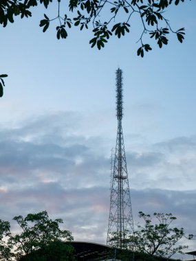 Uzun, metalik kafes iletişim kulesi, çevreleyen ağaç dalları ve yaprakları, teknoloji ve doğa çatışmaları ile dramatik bir akşam gökyüzüne ev sahipliği yapıyor..