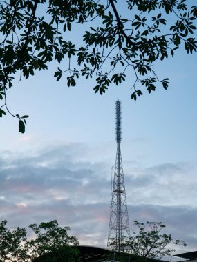 Uzun, metalik kafes iletişim kulesi, çevreleyen ağaç dalları ve yaprakları, teknoloji ve doğa çatışmaları ile dramatik bir akşam gökyüzüne ev sahipliği yapıyor..