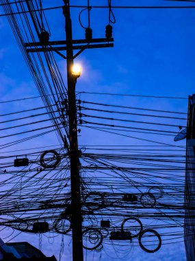 Yoğun elektrik kabloları ve kablolar ahşap bir direğin etrafında yoğun bir siluet oluşturuyor. Derin mavi bir gökyüzüne karşı, kentsel elektrik altyapısı karmaşıklığı..