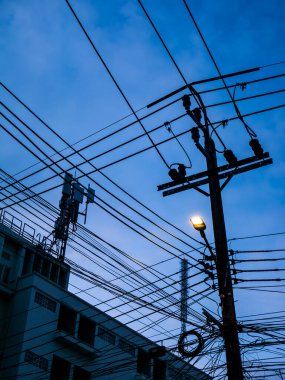 Arka planda haberleşme kuleleri olan bulutlu bir gökyüzüne karşı siluetlenmiş küçük bir ampulle ahşap elektrik direğinin üzerindeki karmaşık elektrik kabloları ve kablolar..