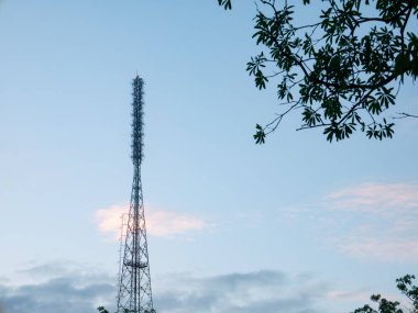 Uzun, metalik kafes iletişim kulesi, çevreleyen ağaç dalları ve yaprakları, teknoloji ve doğa çatışmaları ile dramatik bir akşam gökyüzüne ev sahipliği yapıyor..