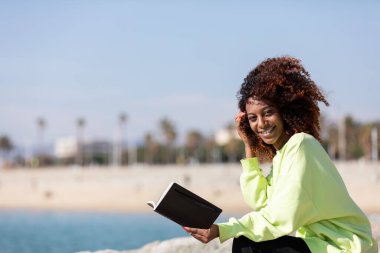 Güneşli bir günde kıyıda oturmuş kitap okuyan gülümseyen Afro-Amerikan kadın.