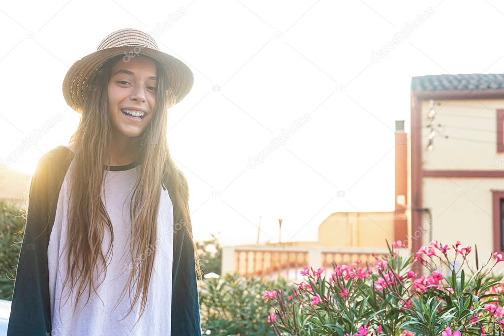 Young happy teenager traveler girl with summer clothes and hat standing in a street outdoors while looking at camera