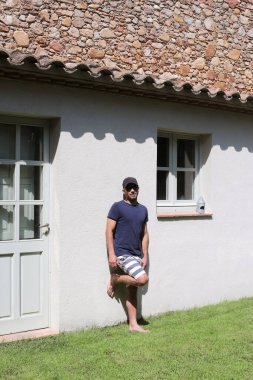 Güneş gözlüklü ve mayosu dışarıda duvara dayanmış bir erkek.