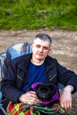 Turist, köpekli siyah pug ile turistik koltukta oturmuş, kareli ekosenin altında, doğal arka planda gülümsüyor. Kapatın. Seçici yumuşak odaklanma. Yüzeyin derinliği. Metin kopyalama alanı.