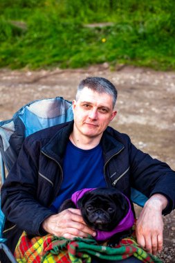 Turist, köpekli siyah pug ile turistik koltukta oturmuş, kareli ekosenin altında, doğal arka planda gülümsüyor. Kapatın. Seçici yumuşak odaklanma. Yüzeyin derinliği. Metin kopyalama alanı.