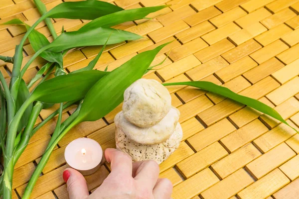 Spa, zen, massage, relaxation concept. Woman hand holding lighting candle, bamboo leaves and white stone pyramid on wooden background. Top view. Flat lay. Close up. Selective focus. Copy space.