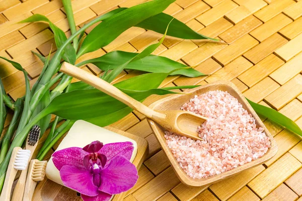 Spa, zen, massage concept. Pink himalayan bath salt with spoon, bamboo leaves, purple orchid, soap in soap dish on wooden carpet background. Top view. Close up. Selective focus. Text copy space.