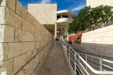 Ziyaretçiler, Los Angeles, Kaliforniya 'daki Getty Center' daki taştan çizgili rampa ve yürüyüş yolu boyunca yürüyorlar. Çevrelerinde travertin duvarlar ve manzaralı yeşillikler bulunuyor.