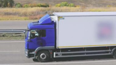 Güneşli bir havada beyaz bir araba otoban boyunca sürer. Otoyolda kamyon var.