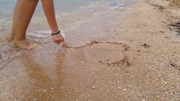 La ola lava el corazón anidado en la arena. La mujer dibuja un corazón en la arena. Una mujer dibuja un corazón en la orilla del mar que es arrastrado por una ola. — Vídeos de Stock