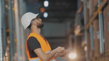 Modern bir depoda bir adam. Depo müdürü depoyu kontrol ediyor. Depoda kask takan bir işçi.