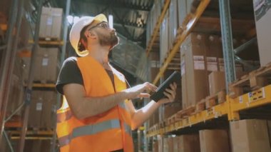 Modern bir depoda bir adam. Depo müdürü depoyu kontrol ediyor. Depoda kask takan bir işçi.