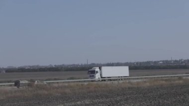 Güneşli bir havada beyaz bir araba otoban boyunca sürer. Otoyolda kamyon var.