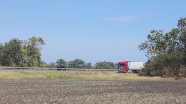Güneşli bir havada beyaz bir araba otoban boyunca sürer. Otoyolda kamyon var.