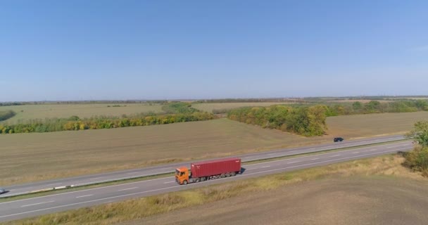 Ciężarówka z czerwonym kontenerem jedzie wzdłuż drogi. Ciężarówka z pomarańczową kabiną transportuje czerwony widok kontenera z drona — Wideo stockowe