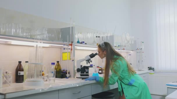 Una chica en el laboratorio. Hermosa chica en el laboratorio mira a través del microscopio — Vídeos de Stock