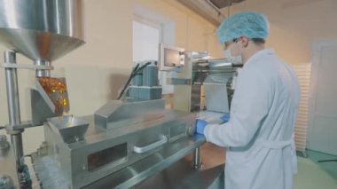 Yüzünde maske olan bir işçi. Jelatin kaplı tabletlerin üretimi için bir mekanizma. Koruyucu maskeli bir adam ve uyuşturucu üretim tesisinde önlük giyen bir adam..