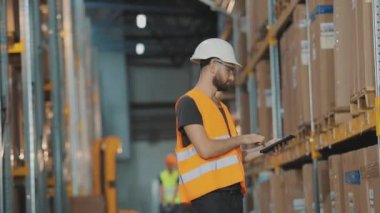 Modern bir depoda bir adam. Depo müdürü depoyu kontrol ediyor. Depoda kask takan bir işçi.
