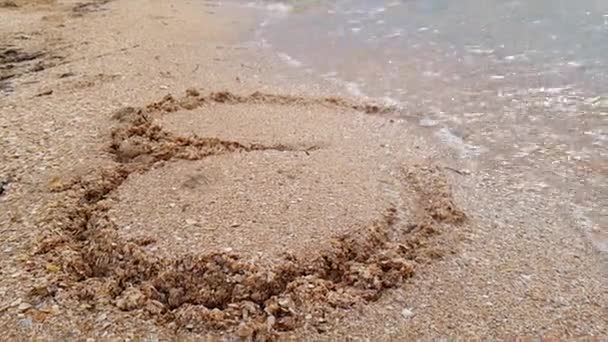 Женщина рисует сердце на берегу моря, которое смывает волна. Волна смывает сердце, спрятанное в песке. Женщина рисует сердце в песке — стоковое видео