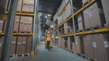 Depoda forklift kullanan bir işçi. İşçi depoya doğru yürür. Depoda çalışan bir işçi elektrikli forklift taşır.