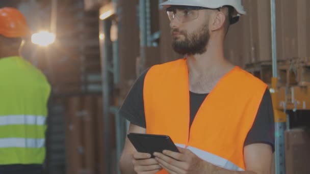 Aktive Arbeit in einem Lager. Das Lager einer modernen Fabrik. Ein Lagerarbeiter mit Helm inspiziert die Ware — Stockvideo