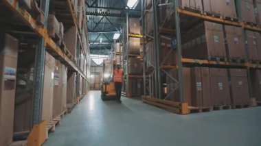 Depoda forklift kullanan bir işçi. İşçi depoya doğru yürür. Depoda çalışan bir işçi elektrikli forklift taşır.